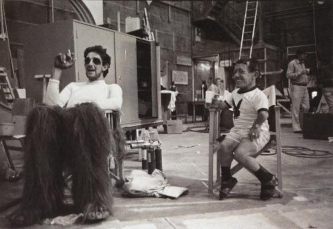 Two men sit on set in a backstage area. One wears furry costume legs, and the other has a shirt with a Playboy bunny logo. They're relaxed, surrounded by set equipment, ladders, and crew in the background.