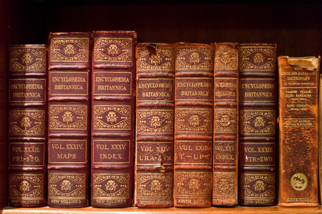 A row of old, worn Encyclopedia Britannica volumes and a New English-Japanese Dictionary with gold-embossed spines are arranged upright on a wooden bookshelf.