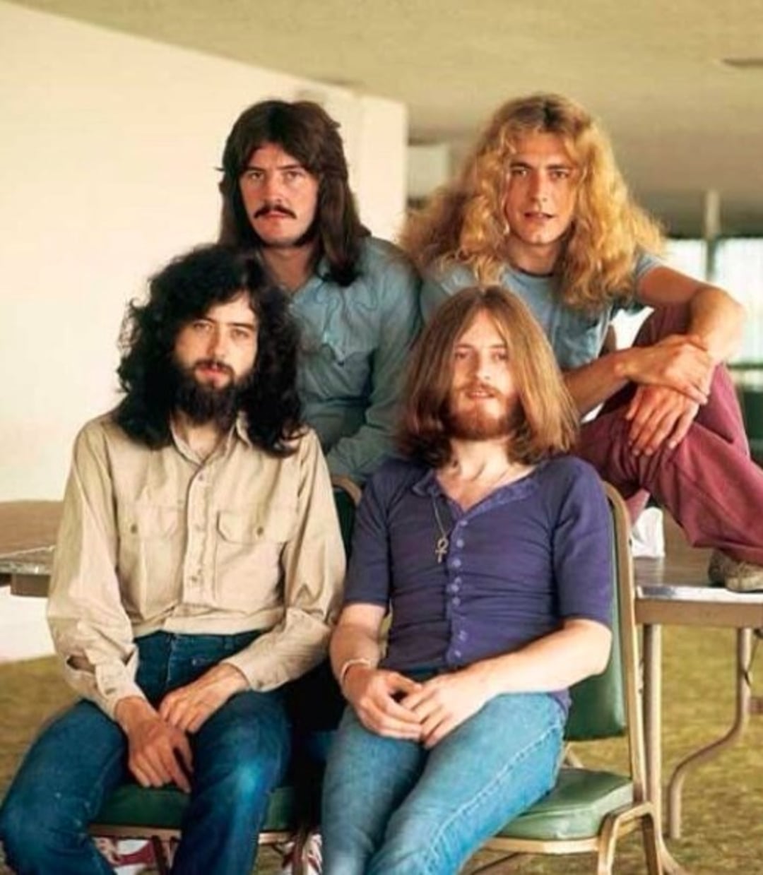 Four men with long hair and casual clothing pose together indoors; two stand in back, two sit in front, all looking at the camera with relaxed expressions in a brightly lit room.