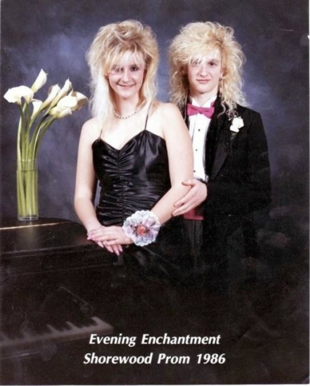Two teens with teased blonde hair pose for a formal prom photo. The girl wears a black dress and corsage, and the boy wears a tux with a pink bow tie. White lilies in a vase sit on a piano. Text reads “Shorewood Prom 1986.”
