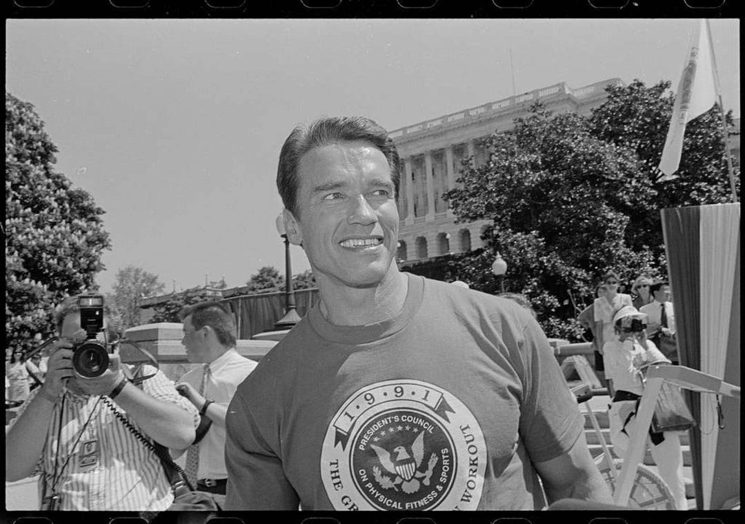 A smiling man in a t-shirt with a fitness emblem stands outdoors near a government building, surrounded by people, including a photographer capturing the scene. Trees and exercise equipment are visible in the background.