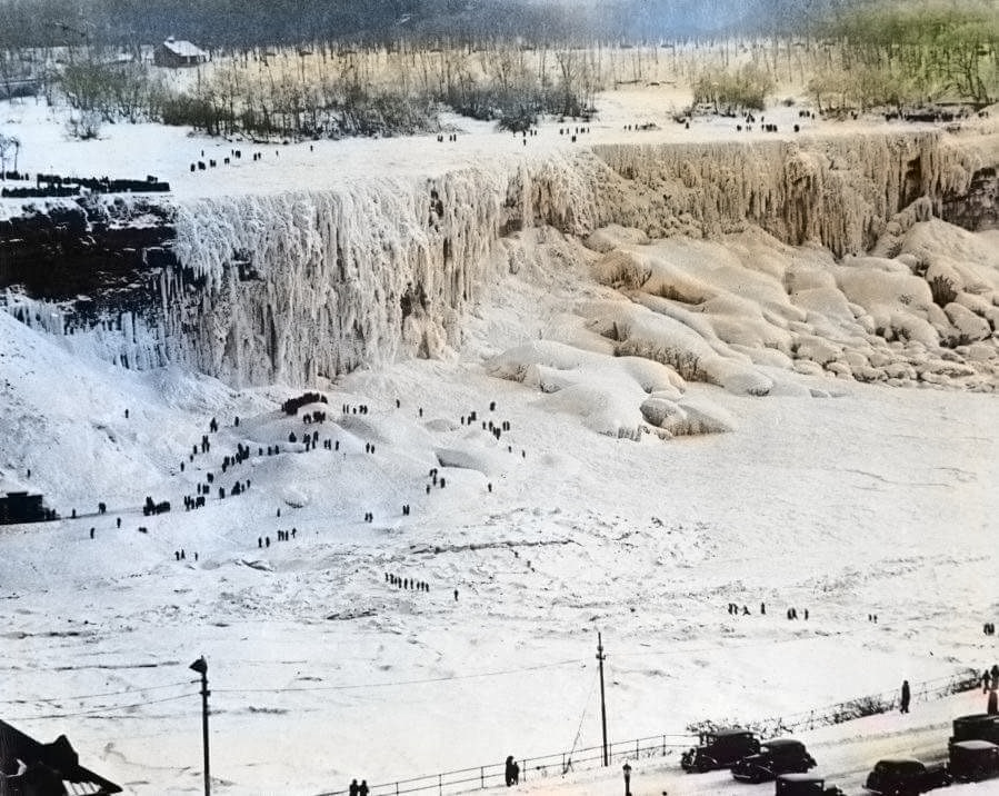 A wide view of a frozen waterfall and river, with people walking across the icy landscape. Snow and ice cover the cliffs and ground, while scattered trees and cars appear in the distance.