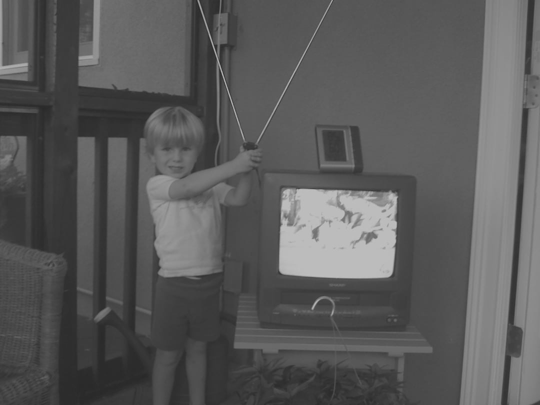 A young child stands indoors holding TV antenna wires above an old box-style television displaying a blurry image. The TV sits on a small table next to a wicker chair and a wall-mounted device. The scene appears vintage.