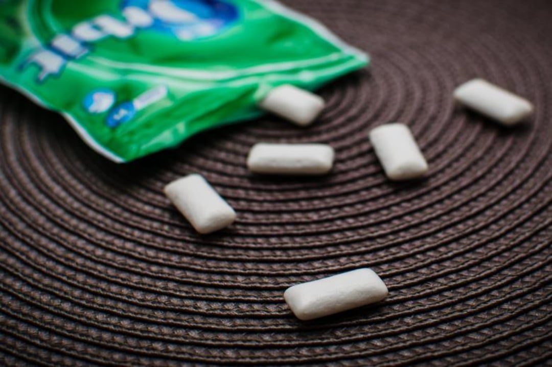 A green pack of chewing gum is open on a brown woven surface, with several white rectangular gum pieces scattered nearby.