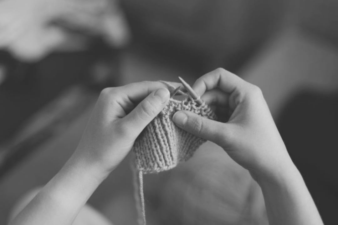 Close-up of two hands holding knitting needles, working on a knitted piece of fabric. The image is in black and white, with a blurred background.
