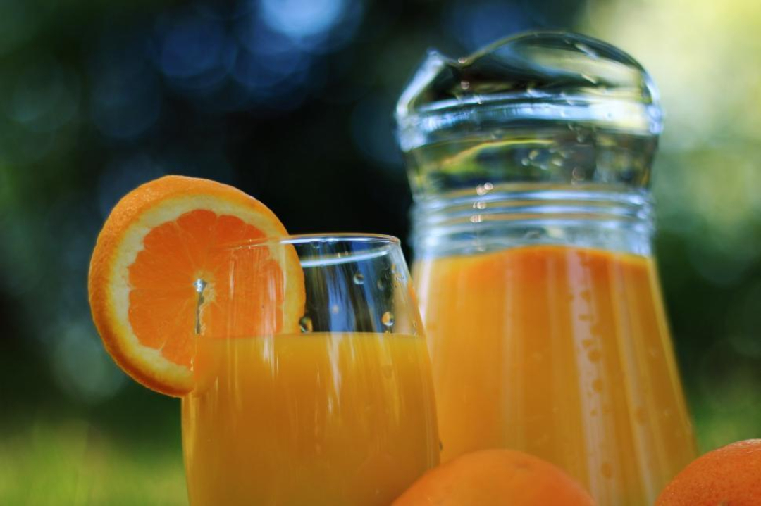 A glass and a pitcher of orange juice are placed outdoors. The glass has a slice of orange on its rim, and whole oranges are visible in the foreground. The background is blurred greenery.