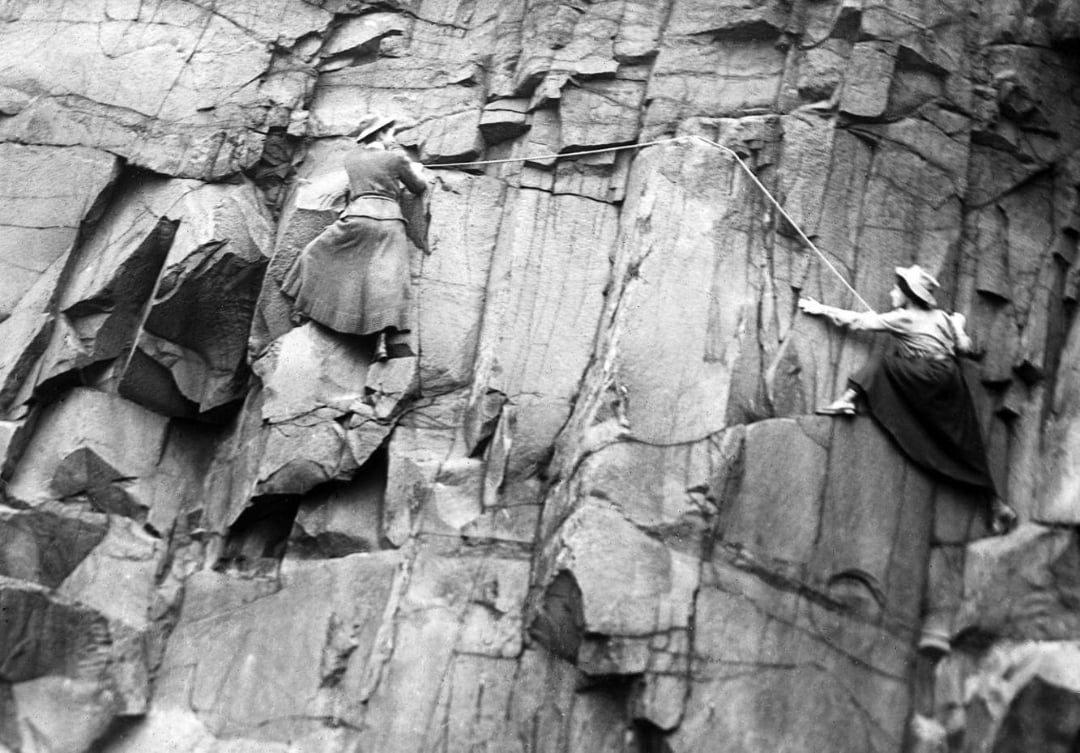 Two women in skirts and hats climb a steep, rocky cliff, using ropes for support. The black-and-white photo has a historical feel, highlighting early rock climbing.
