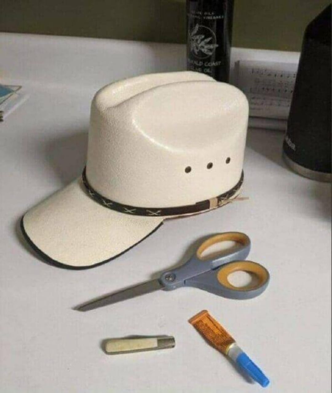 A small white cowboy hat made from a disposable cup sits on a desk, surrounded by scissors, a tube of glue, and a utility knife, suggesting it was crafted from nearby materials.