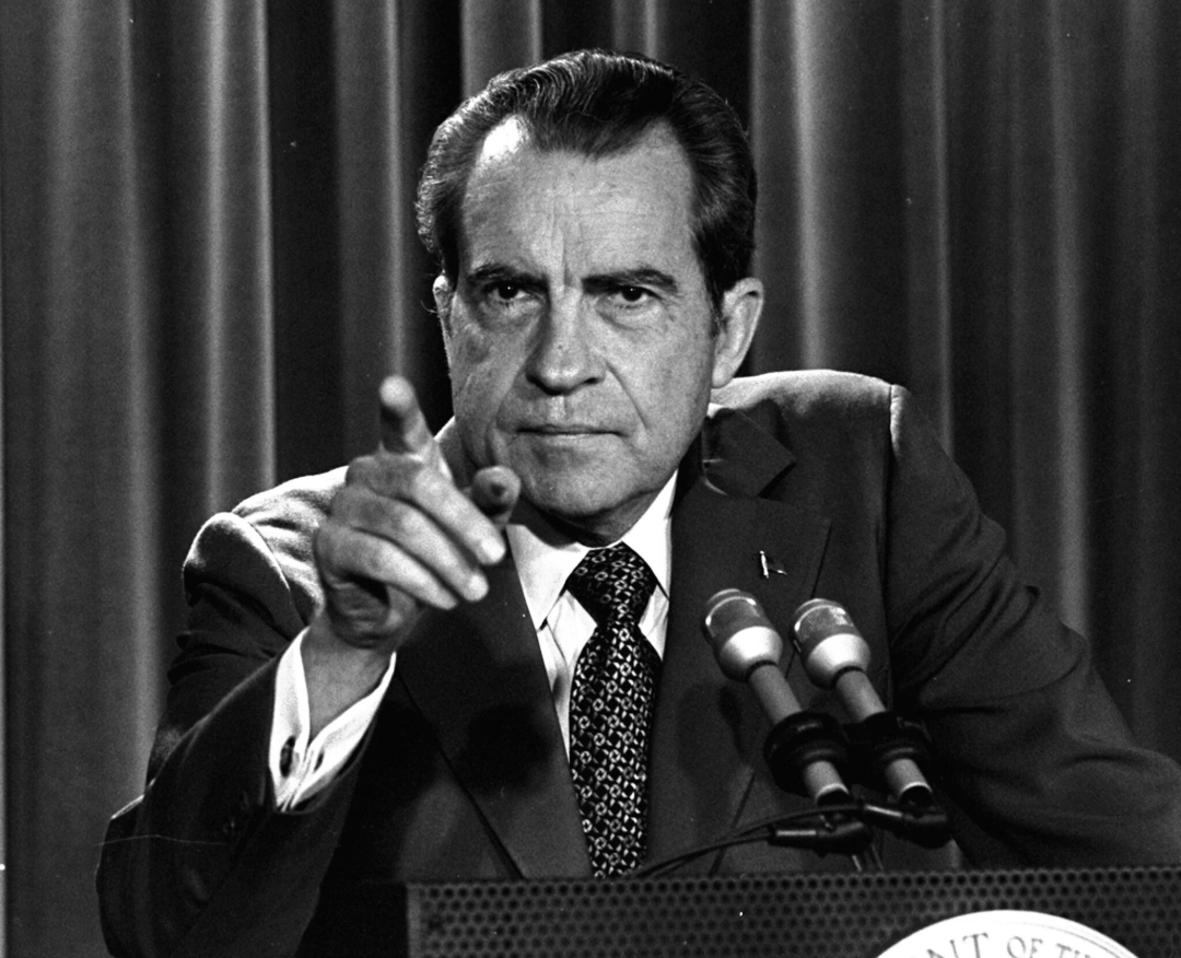 A man in a suit and tie sits at a podium with microphones, pointing forward and looking serious. There is a curtain in the background.