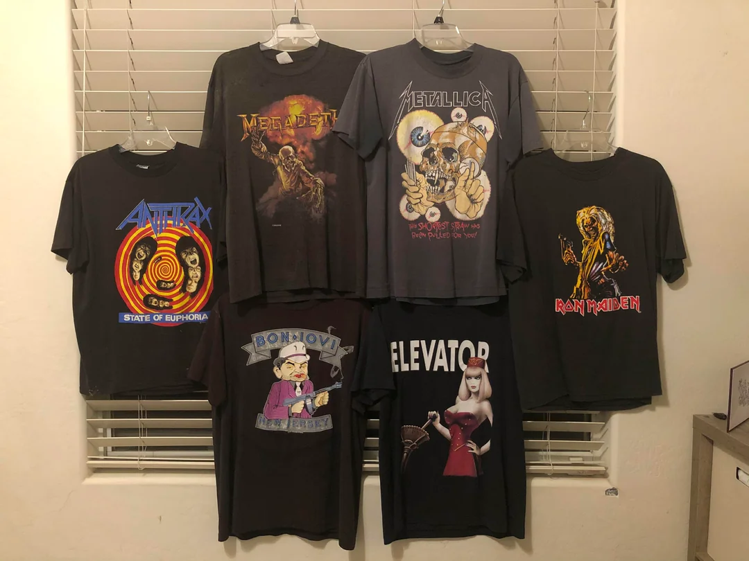 Six graphic t-shirts hang in front of window blinds, featuring designs from Anthrax, Megadeth, Metallica, Iron Maiden, Bon Jovi, and a shirt with the word "ELEVATOR" and an animated character.