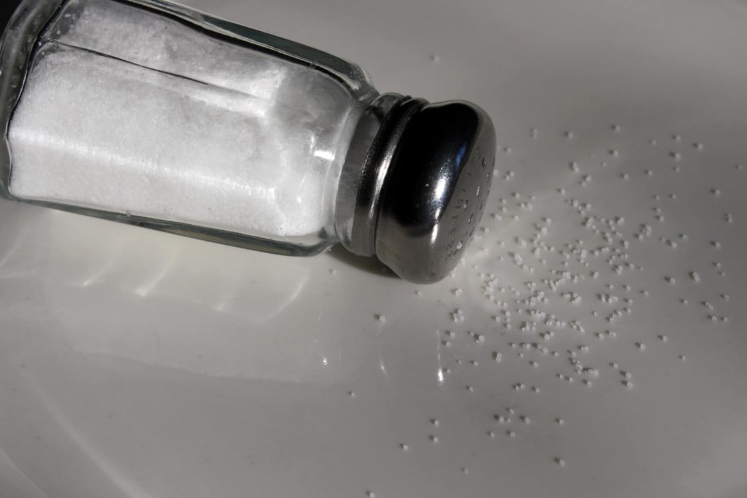 A salt shaker lies on its side on a white surface, with salt granules spilled out next to it.