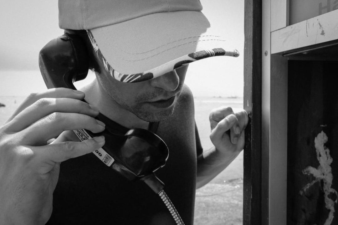 A person wearing a cap is talking on a payphone, holding the receiver to their ear and standing partially inside the phone booth. The photo is in black and white.