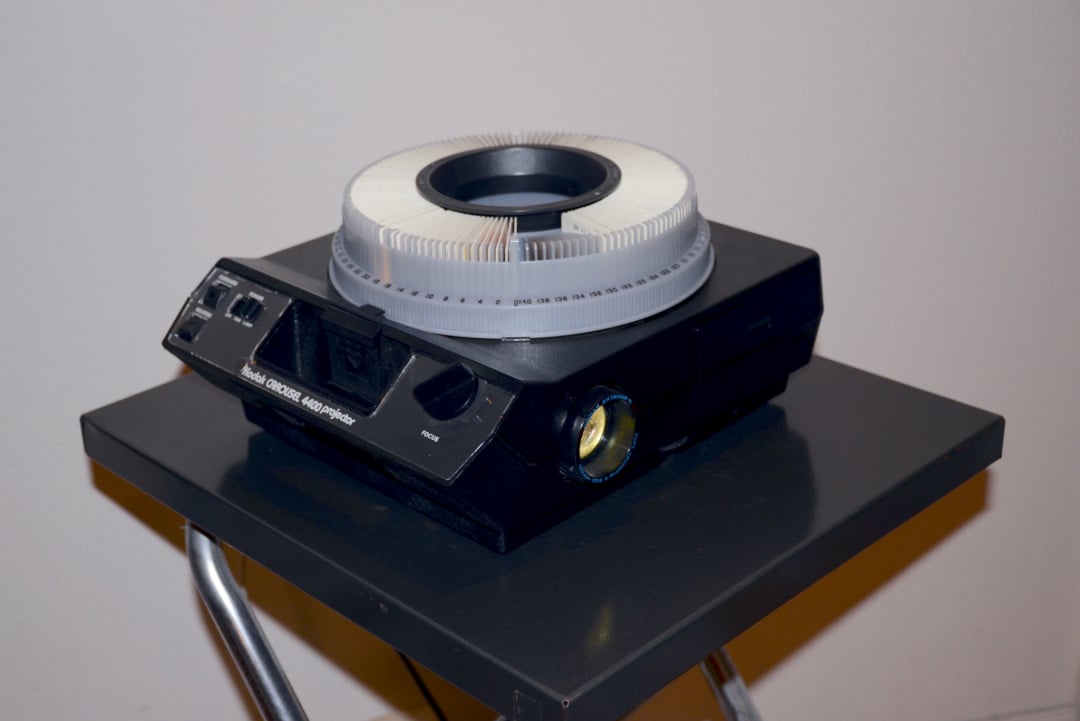 A Kodak Carousel 4600 slide projector sits on a metal stand, with a circular tray of slides loaded on top, against a plain beige wall.