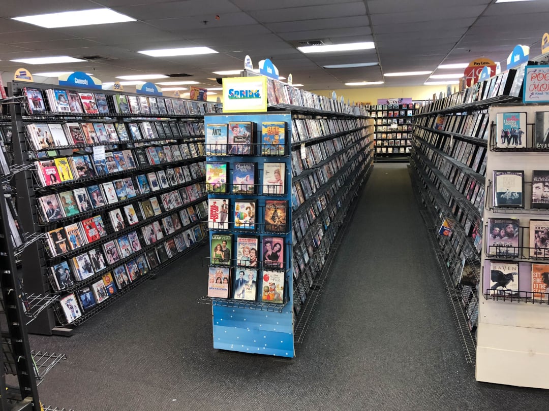 Aisles of DVDs and Blu-rays line the shelves in a well-lit video rental store, with colorful covers facing outward and a blue display stand in the center of the image.