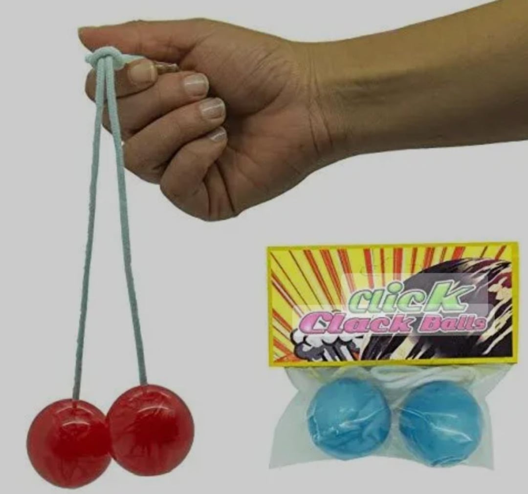 A hand holds two red plastic balls connected by a string. Next to it is a package labeled "Click Clack Balls" containing two blue balls connected by a string.