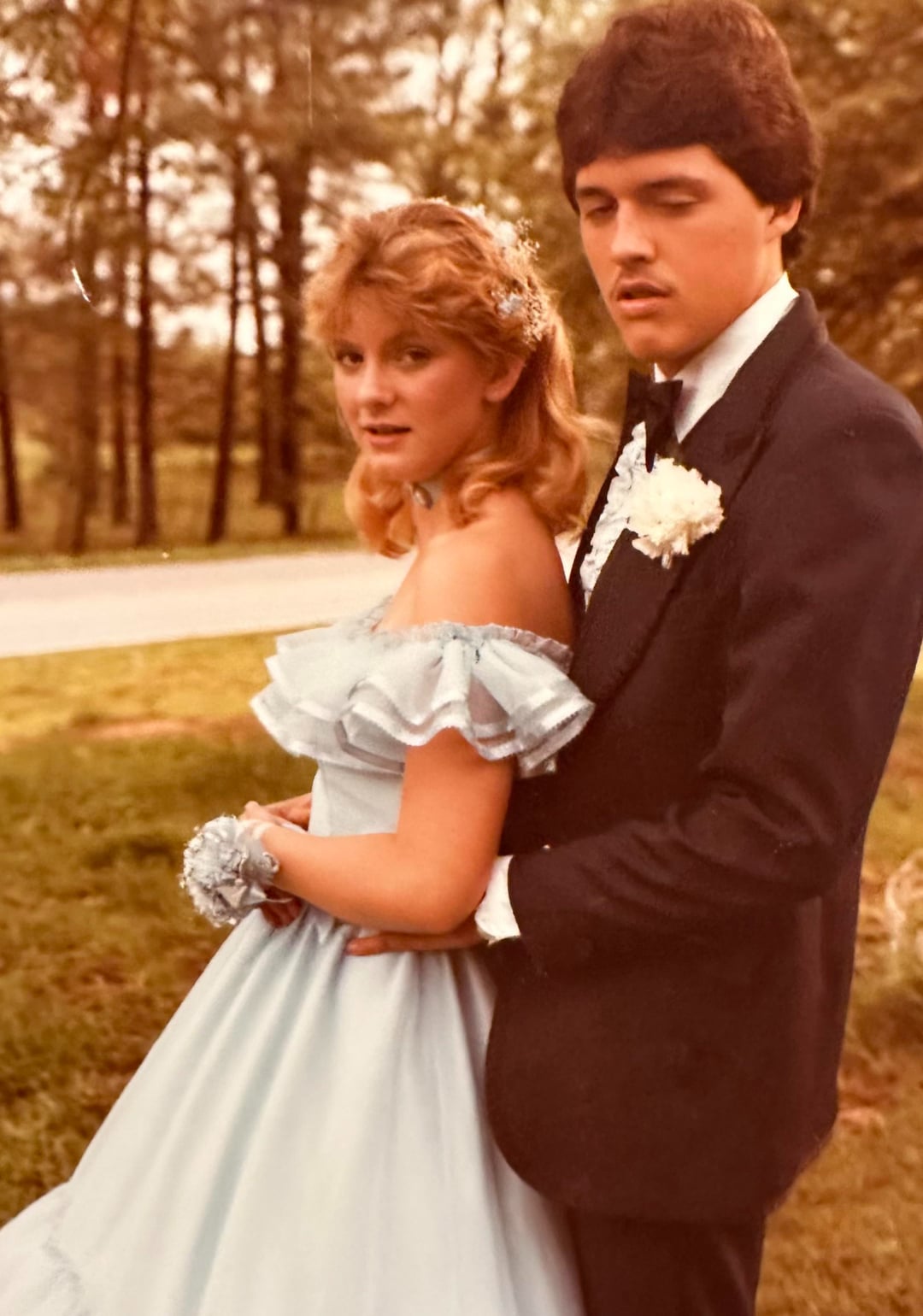 A young couple in formal attire pose outdoors. The woman wears a light blue off-the-shoulder gown and corsage, while the man wears a dark tuxedo with a white boutonniere. Trees and grass are visible in the background.