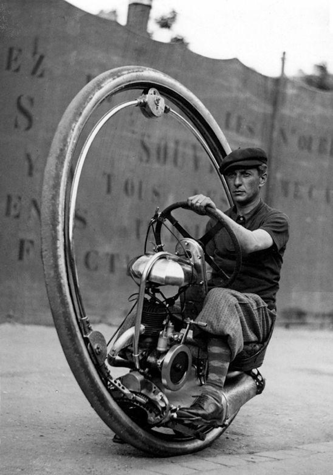 A man wearing a cap and knickerbocker pants rides a vintage monowheel motorcycle, with a large single wheel encircling him and his machine. The background features a faded, painted wall with partially visible text.