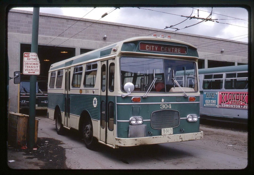 A vintage green and white bus labeled "CITY CENTRE" and numbered 304 is parked outside a garage. The bus is slightly weathered, and another bus is visible in the background.