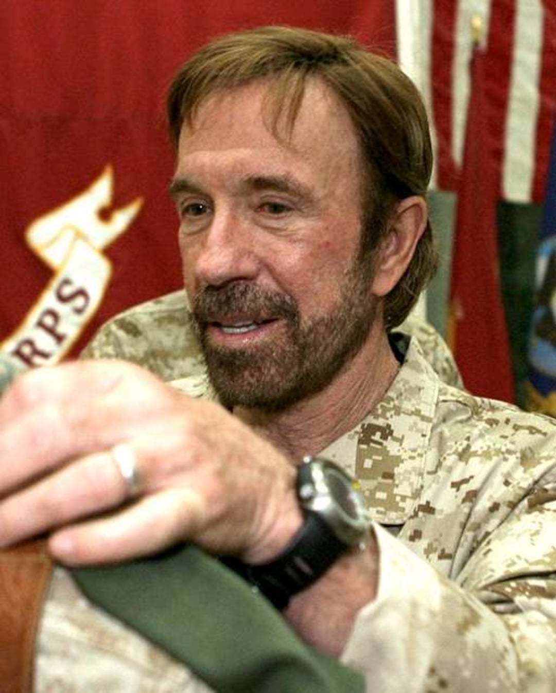 A man with a beard and mustache, wearing military camouflage, holds a green piece of fabric. American flags are visible in the background. The man is looking down and smiling slightly.
