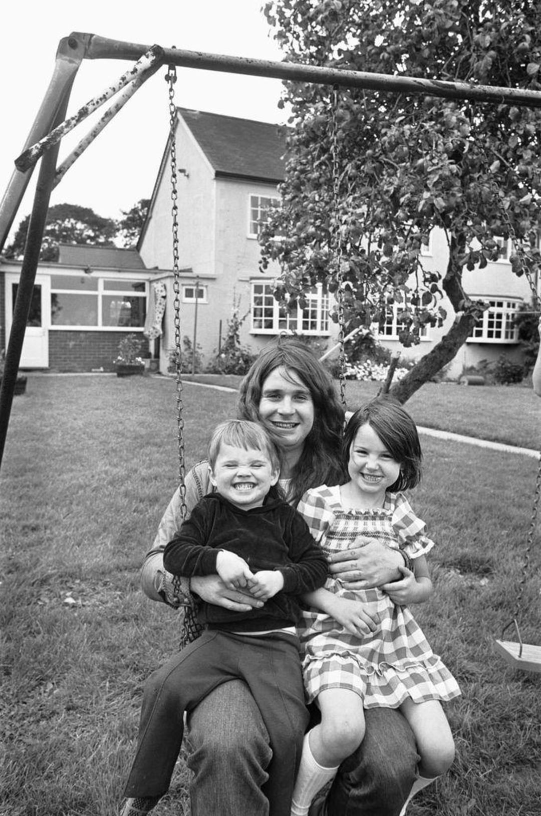 A smiling adult sits on a garden swing holding two happy young children, one in a checkered dress and one in dark clothes, with a house and tree visible in the background.