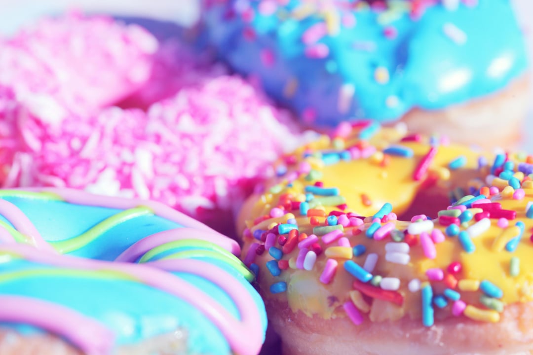 Four colorful donuts topped with bright icing and rainbow sprinkles are arranged closely together. The donuts have blue, pink, and yellow frosting with various decorative toppings.