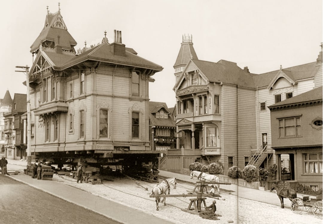 A large Victorian house is being moved down a street using wooden beams and horse-drawn equipment, with similar houses in the background on a cobblestone road. The image is sepia-toned and historic.