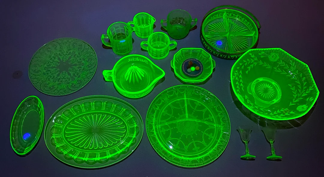 A collection of vintage uranium glass dishes, cups, and bowls glowing bright green under blacklight on a dark surface. The glassware features intricate patterns and various shapes and sizes.