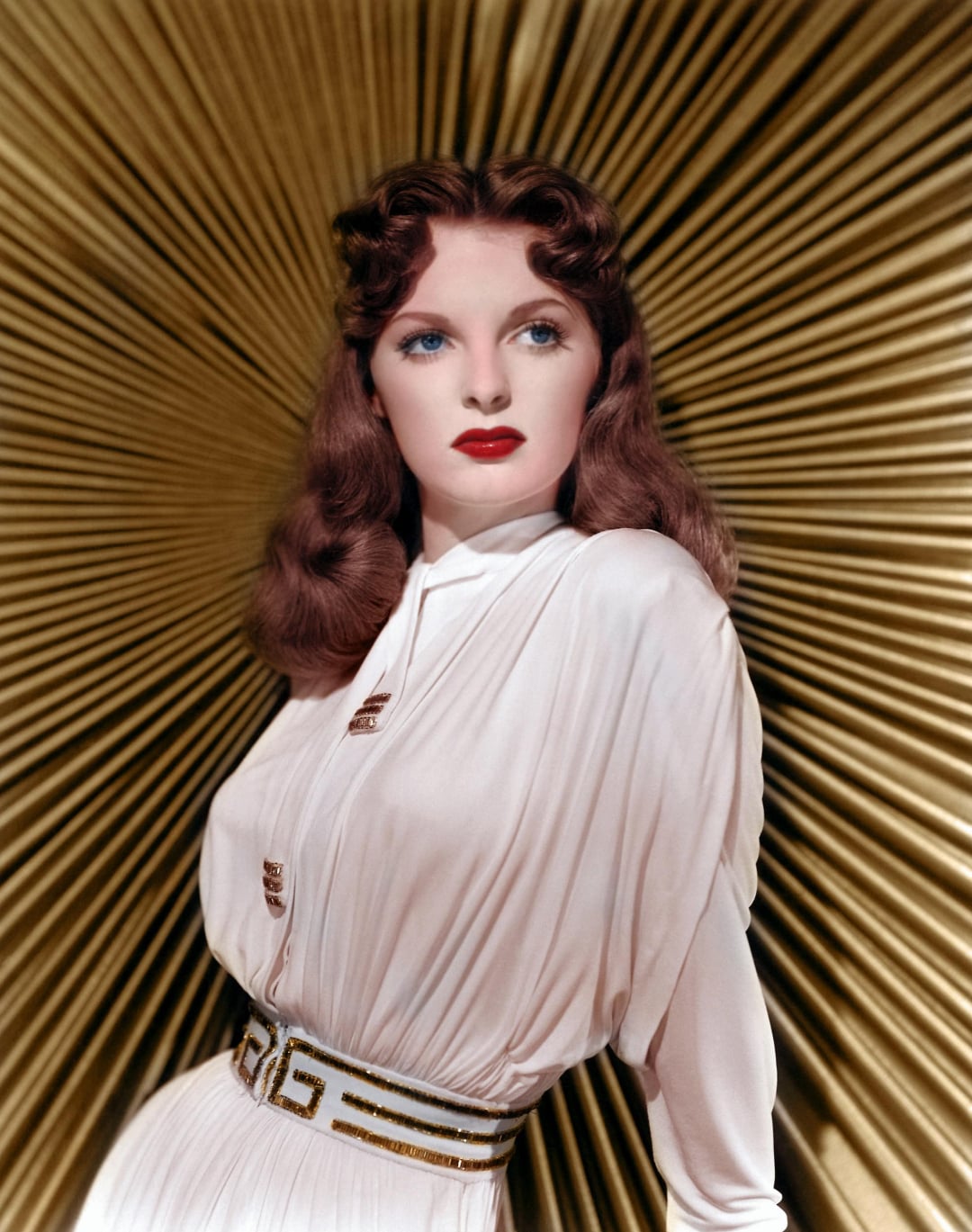A woman with wavy auburn hair and red lipstick wears a white, belted dress. She stands in front of a golden, radiating background, looking slightly to the side with a serene expression.