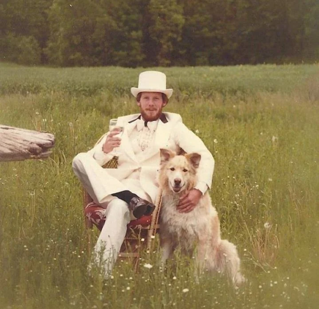 A man dressed in a white suit and top hat sits in a field on a chair, holding a drink and resting his hand on a large, fluffy dog sitting beside him with greenery in the background.
