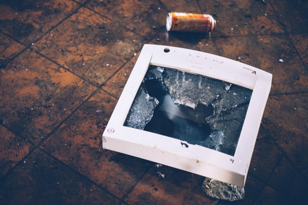 A broken computer monitor with shattered glass lies on a dirty tiled floor next to a crushed soda can.