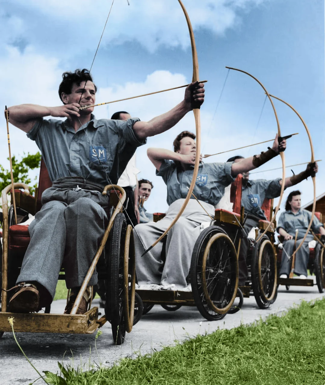 A group of men in wheelchairs, wearing matching uniforms, practice archery outdoors on a sunny day, drawing their bows in unison under a blue sky.