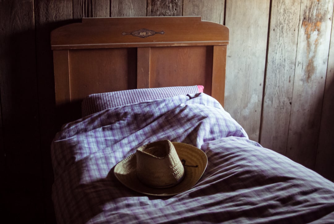 A straw hat rests on a neatly made bed with purple checkered bedding in a rustic wooden room, lit softly by natural light.