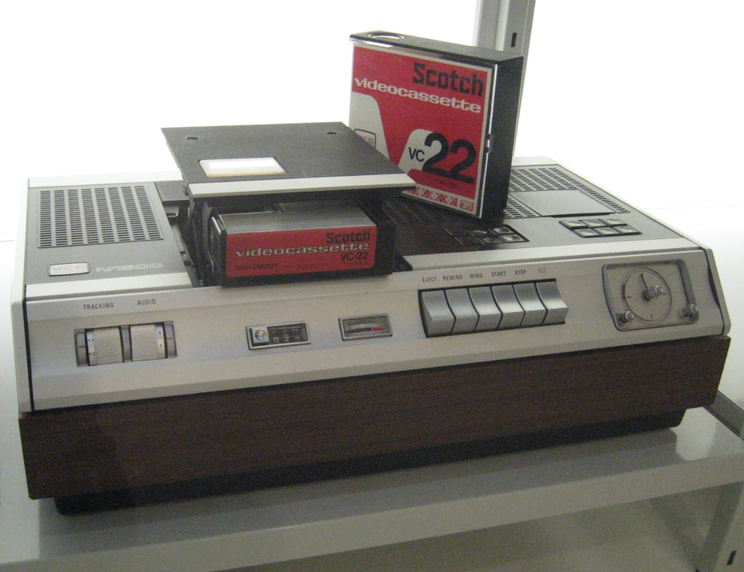 A vintage video cassette recorder with a wood finish sits on a shelf. Its top is open, revealing a Scotch videocassette VC 22 being inserted, and another cassette stands upright next to it.
