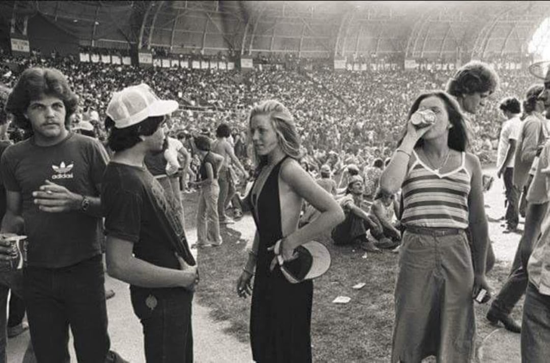 A black-and-white photo of young people at an outdoor concert or festival. Four people stand in the foreground talking and drinking, with a large crowd seated in the background inside a stadium.