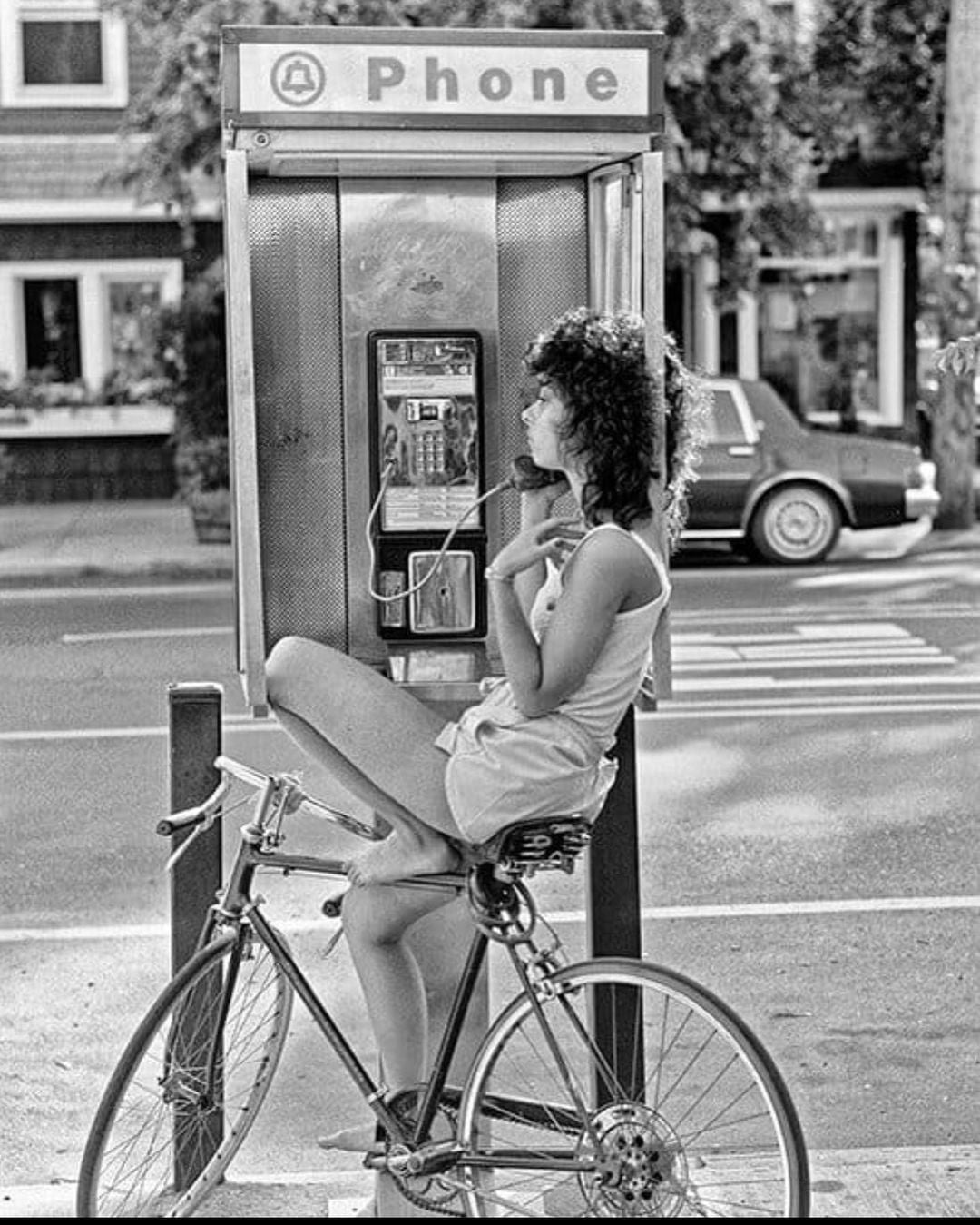 A woman sits on a bicycle, holding a phone receiver at a public phone booth, with one leg propped on the bike. She looks thoughtful, and a car and houses are visible in the background.