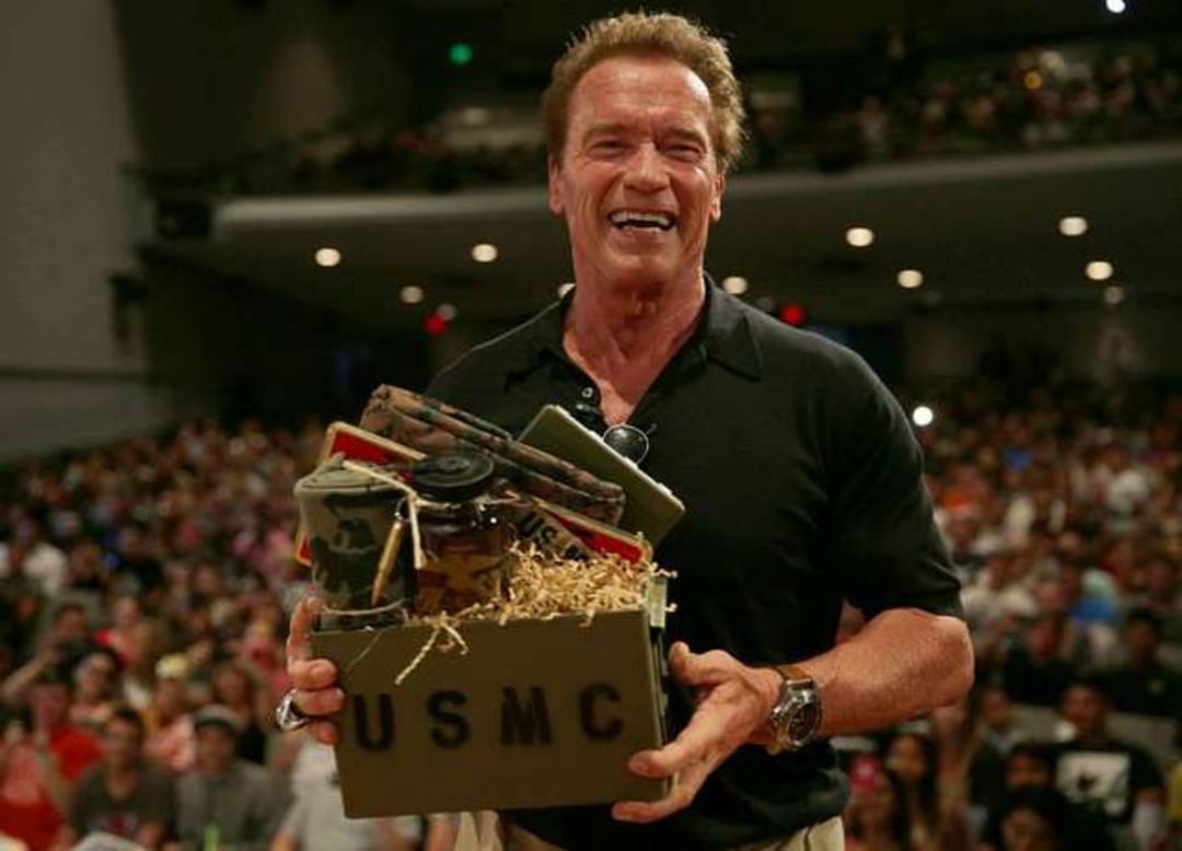 A smiling man in a black polo shirt holds a USMC-themed gift box filled with military items, standing in front of a large audience in an auditorium.