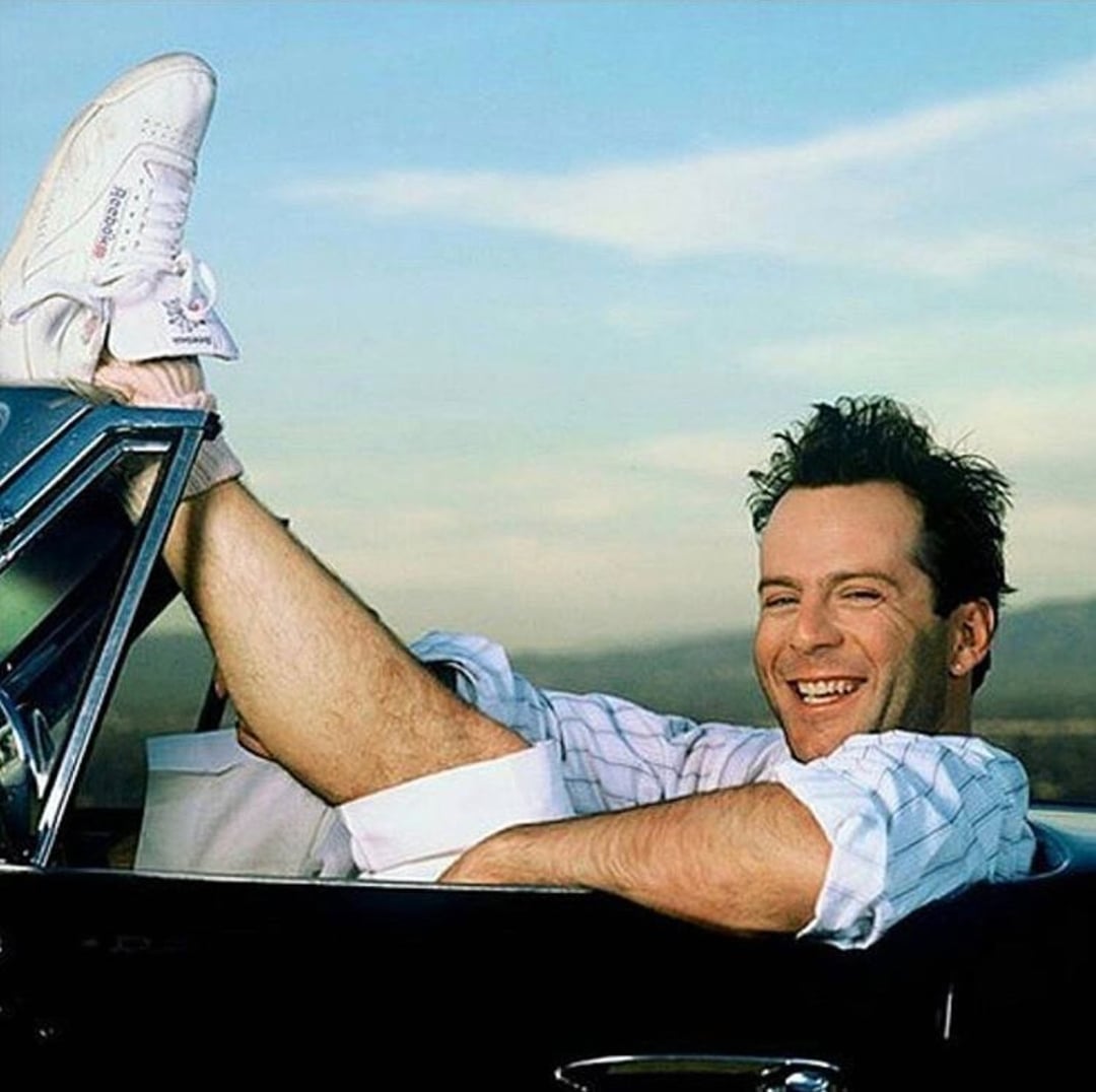 A man with short dark hair sits in a convertible car, smiling with one leg casually propped up on the car door. He wears a white outfit and white sneakers. The background shows a clear blue sky and distant mountains.