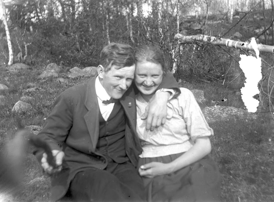 A black-and-white photo of a man in a suit and a woman in a light blouse sitting on grass outdoors. The man has his arm around the woman and both are smiling, with trees and rocks in the background.