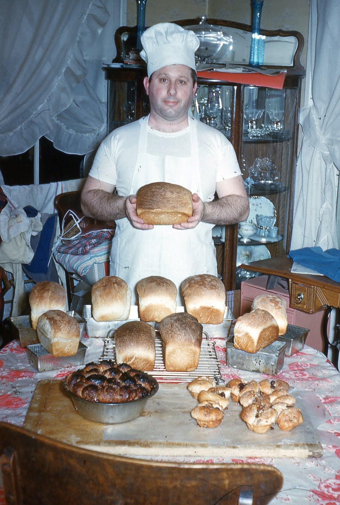 A baker wearing a white hat and shirt proudly holds a loaf of bread in a kitchen filled with various loaves of bread, buns, and pastries displayed on a table. Behind him is a wooden cabinet with glassware.