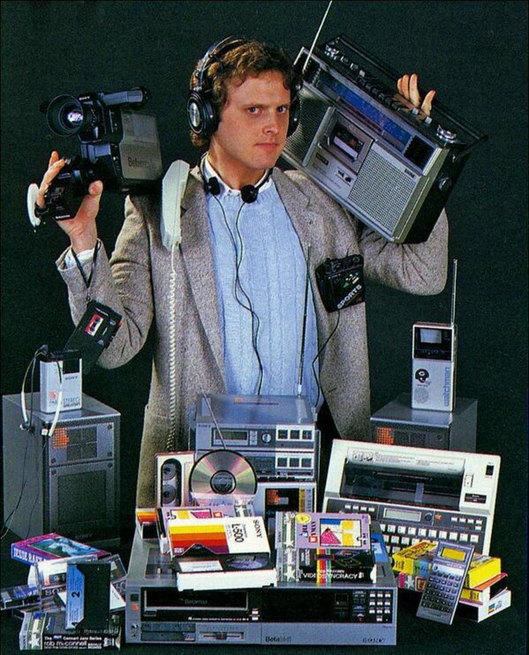 A man in a gray blazer holds a large vintage video camera and a boombox, surrounded by 1980s electronics like cassette tapes, VHS tapes, a turntable, headphones, and various audio-video equipment.