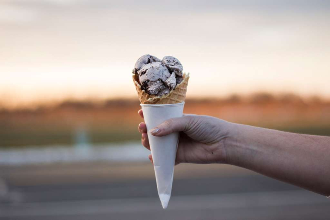 A hand holding an ice cream cone with cookies and cream scoops, wrapped in a paper sleeve, outdoors with a blurred background and soft evening light.
