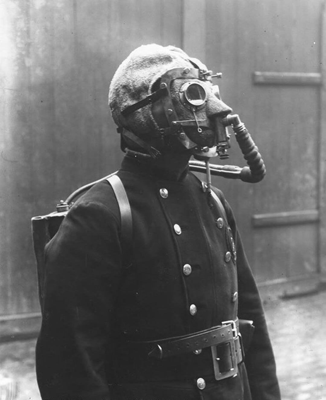 A person wearing an early gas mask and protective uniform, including a helmet and breathing apparatus, stands in front of a large wooden door or gate. The photo is black and white and appears historical.