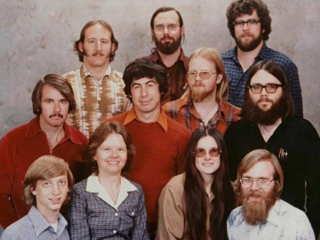 A group of eleven people, ten men and one woman, pose for a portrait in 1970s-style clothing. Most are wearing glasses, and several have long hair or beards. The background is a plain, neutral color.