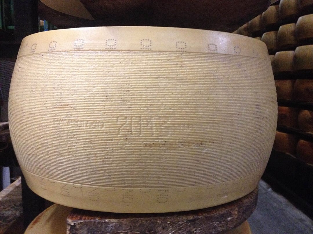 A large wheel of Parmigiano Reggiano cheese sits on a wooden shelf, featuring its signature dotted rind markings and a visible date stamp of 2015.