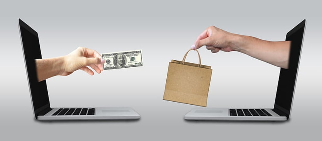 A hand extending a dollar bill from a laptop screen exchanges with another hand offering a small shopping bag from a second laptop, symbolizing online shopping or e-commerce.
