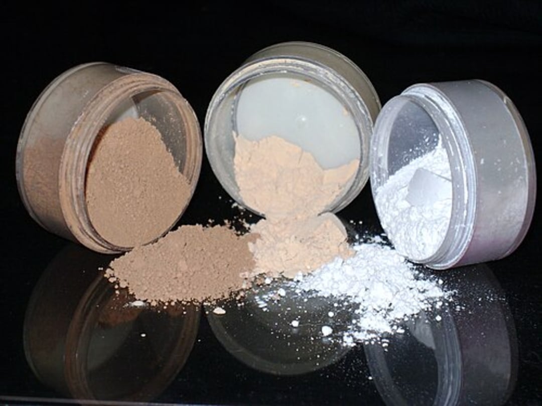 Three open containers of loose powder makeup in brown, beige, and white shades are tipped over, with some powder spilled in front of each on a reflective black surface.