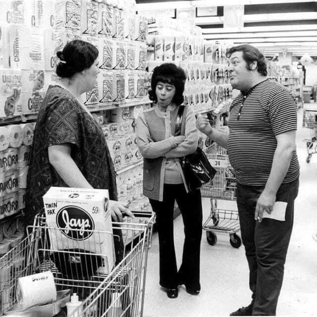 Three people stand in a grocery store aisle filled with paper goods. Two women with shopping carts listen to a man in a striped shirt who is gesturing and holding a paper. The atmosphere seems serious or tense.