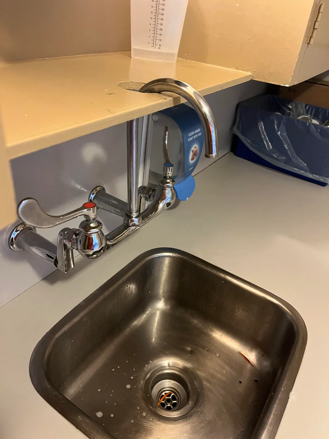 A metal sink with a faucet mounted above a counter, but the faucet is installed on the counter rather than over the sink, making it unusable. A blue trash bin with a liner is visible in the background.
