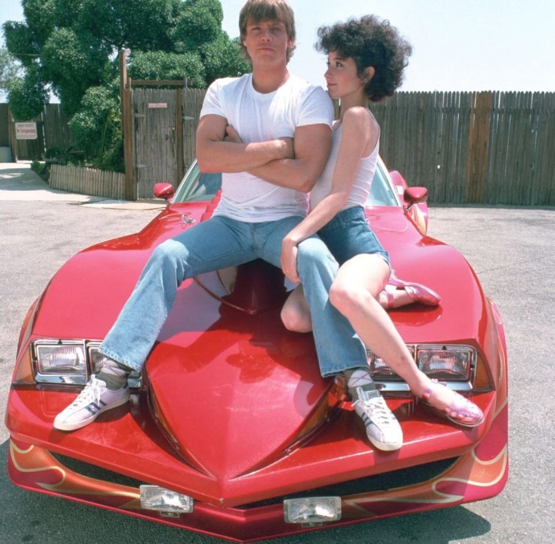 A man and a woman sit on the hood of a shiny red sports car with flaming decals. The man sits with arms crossed, wearing a white T-shirt and jeans. The woman, in a tank top and shorts, sits beside him, looking at him.