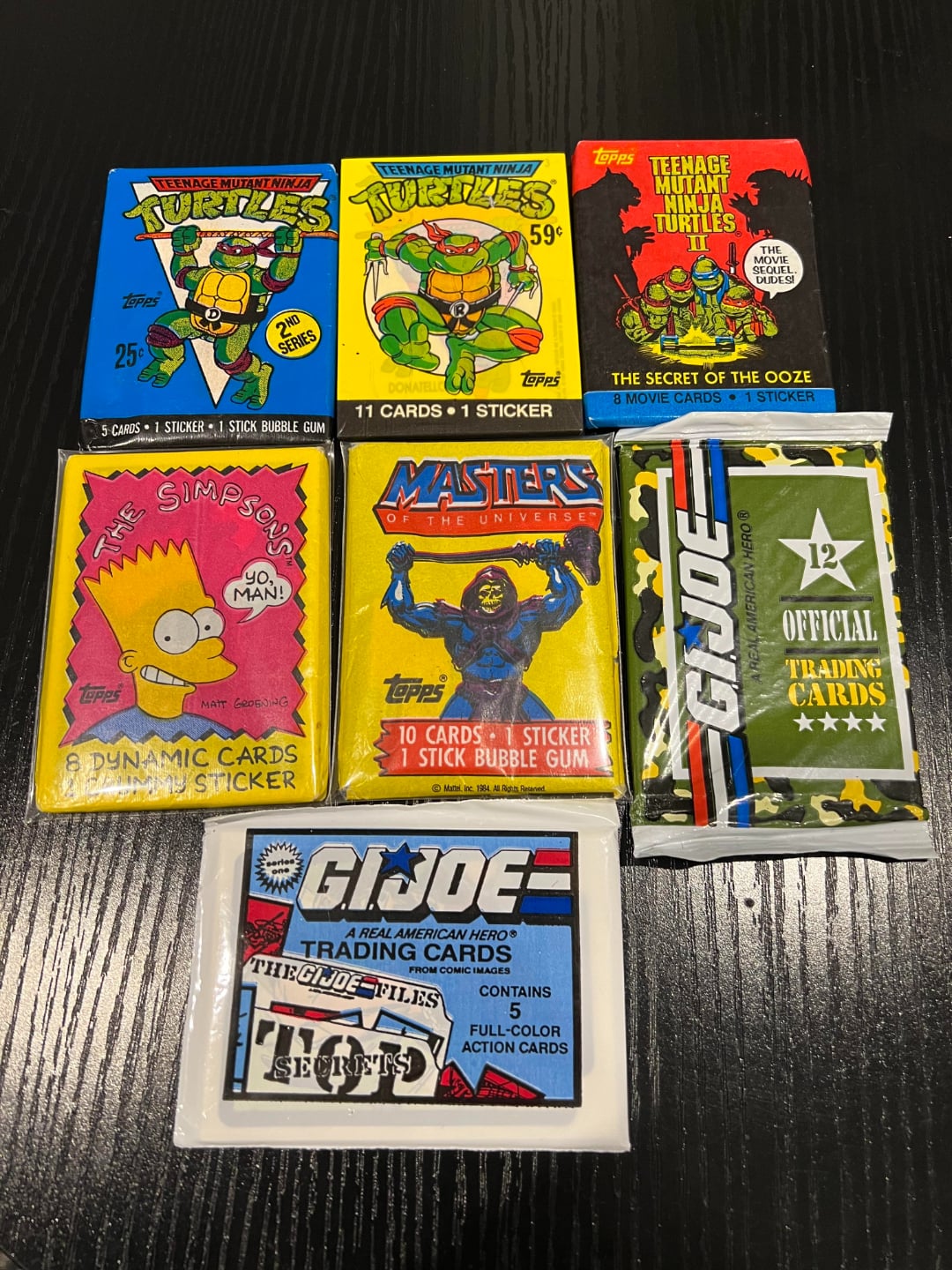 Six packs of vintage trading cards on a black table, featuring designs from Teenage Mutant Ninja Turtles, The Simpsons, Masters of the Universe, and G.I. Joe, each showing colorful artwork and branding.
